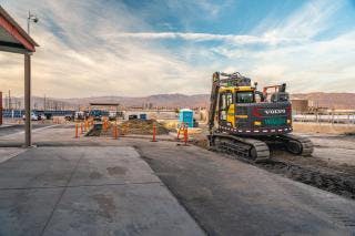 A construction site with an excavator, orange cones, and a porta-potty under a partly cloudy sky.