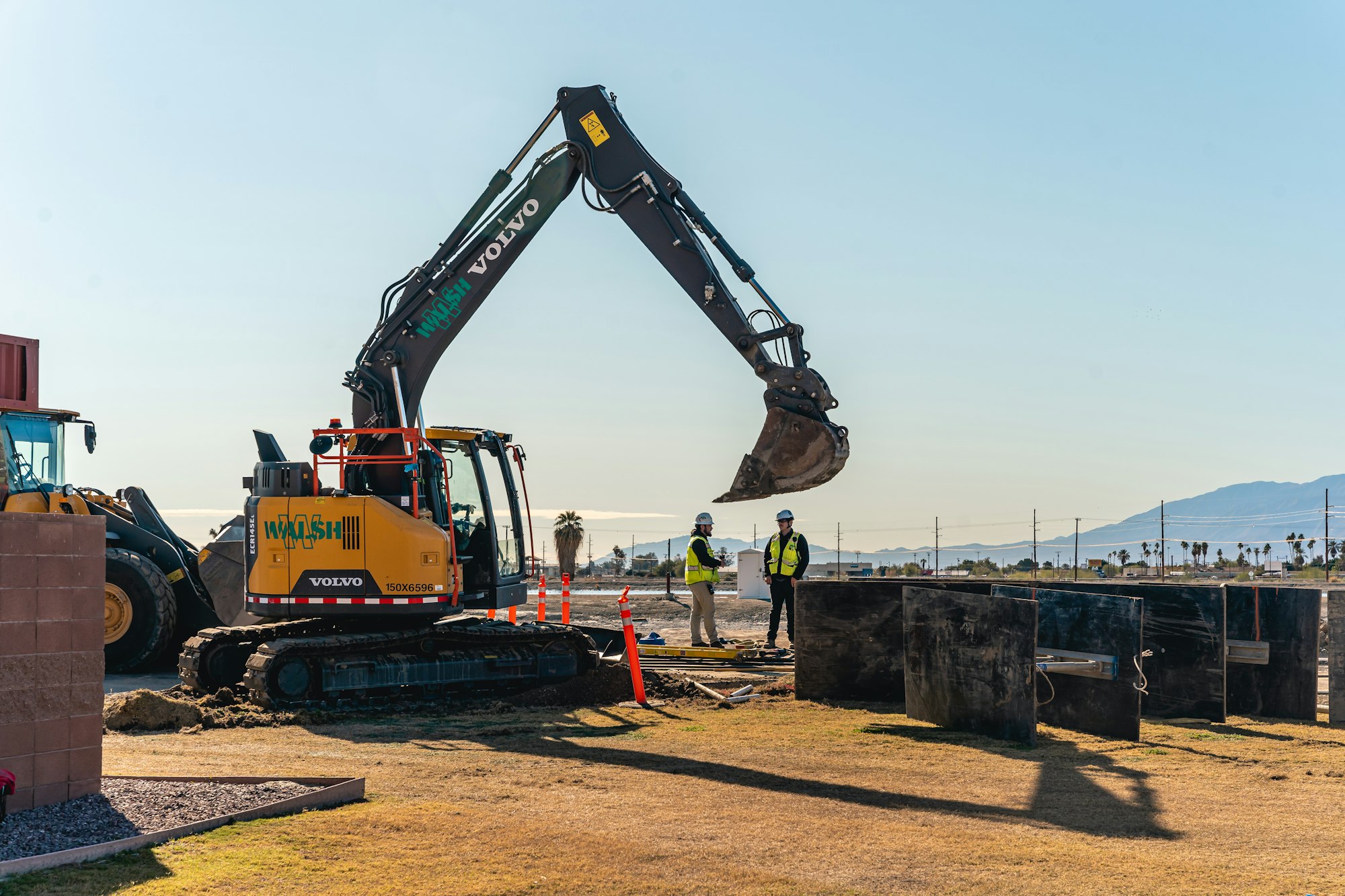 Construction scene with excavator, two workers in safety gear, and barriers in an open area with clear sky and mountains in the background.