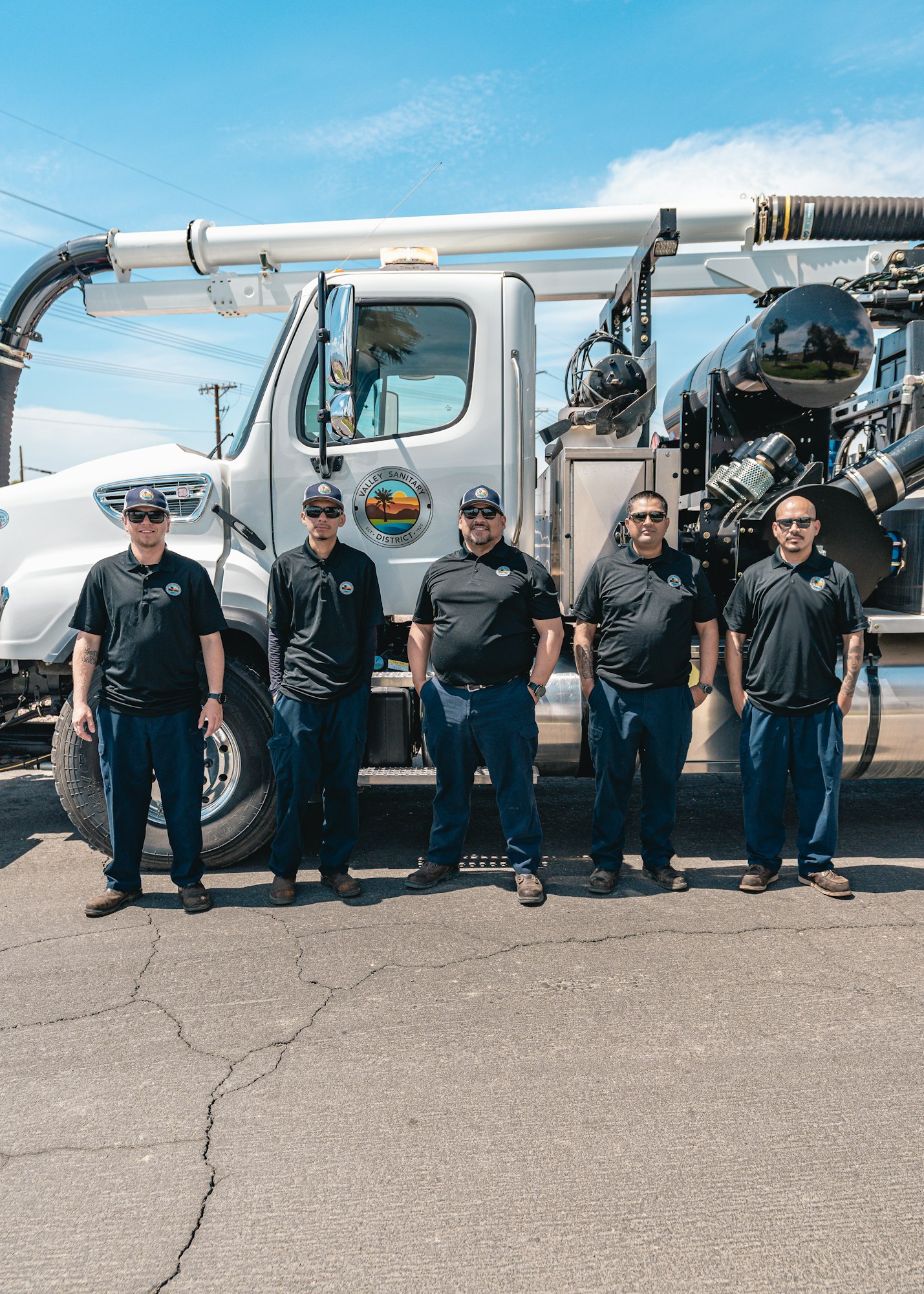 Five people in uniforms stand in front of a large utility truck with a logo, under a clear blue sky.