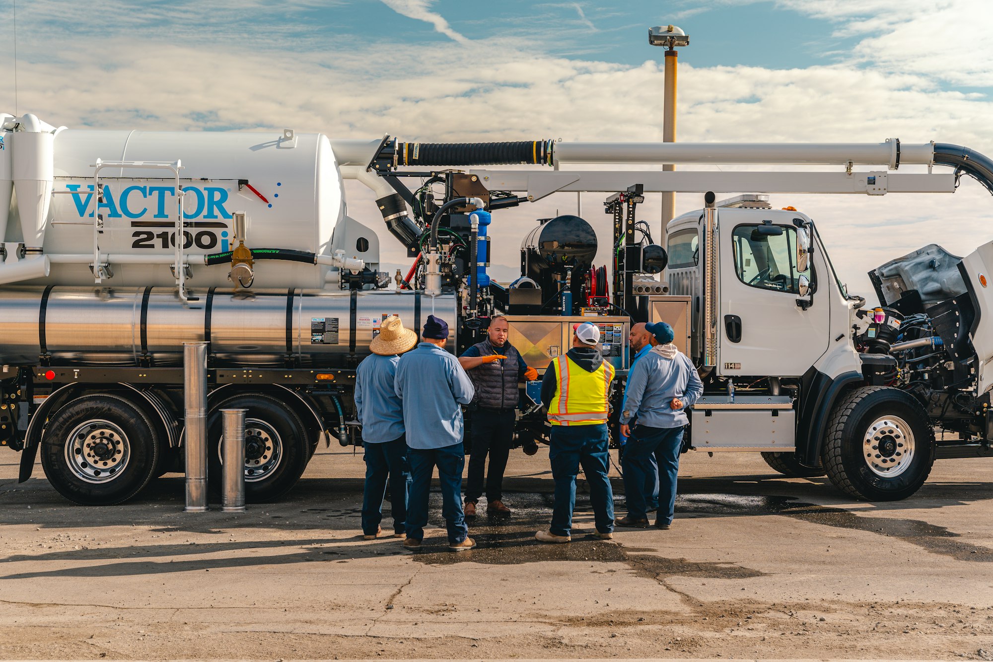 Workers gather around a large utility truck, discussing equipment labeled "VACTOR 2100i" under a partly cloudy sky.