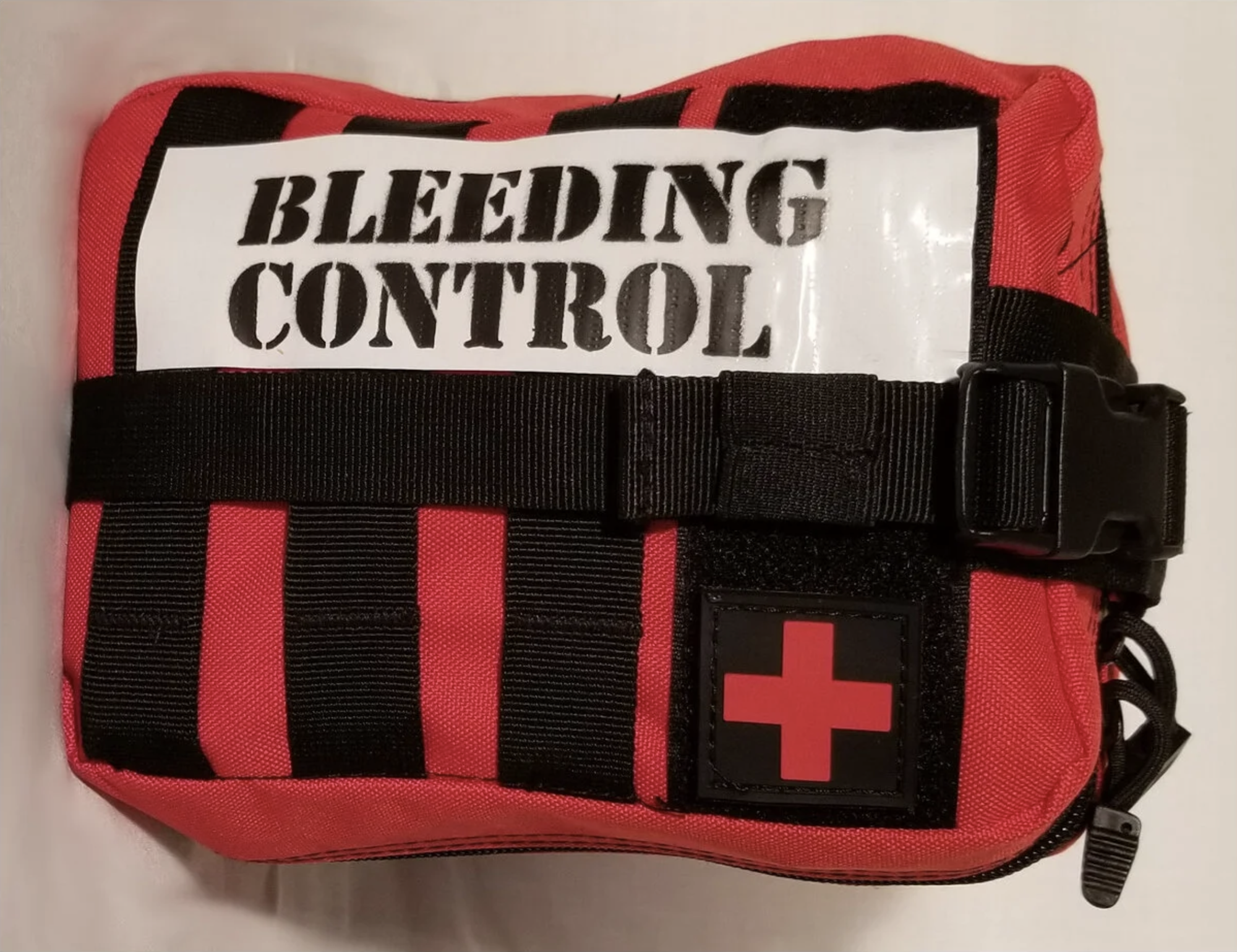 A red first aid kit labeled "Bleeding Control" with a red cross symbol, black straps, and a buckle.