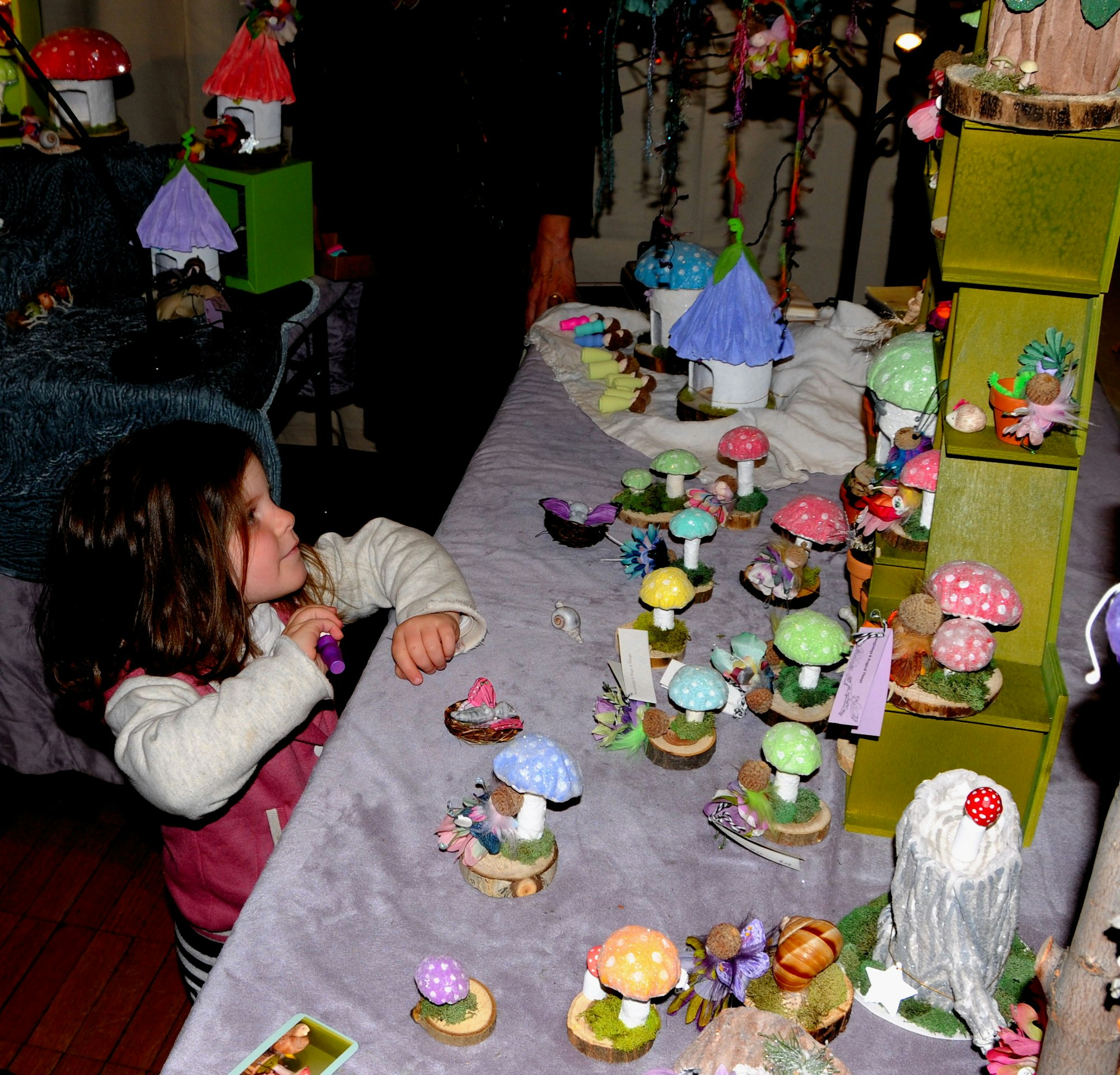A child admires a colorful display of miniature mushrooms and fairy-themed decorations on a table.