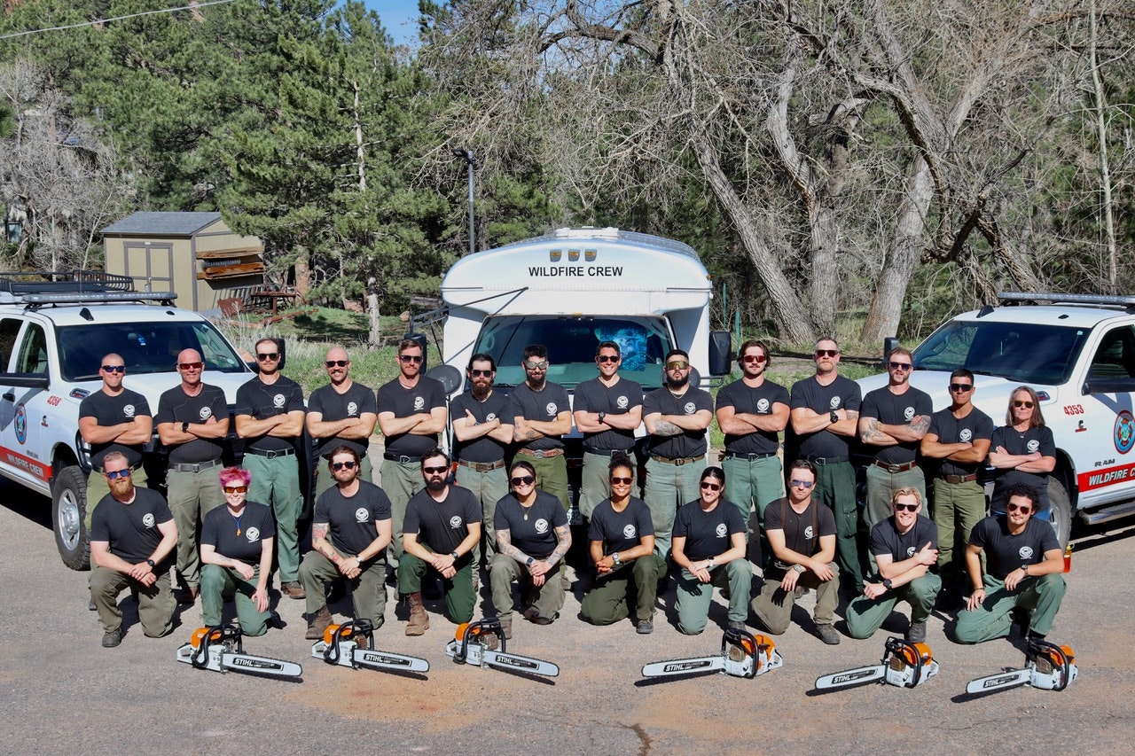 A group of people in matching uniforms poses with chainsaws in front of a wildfire crew vehicle.