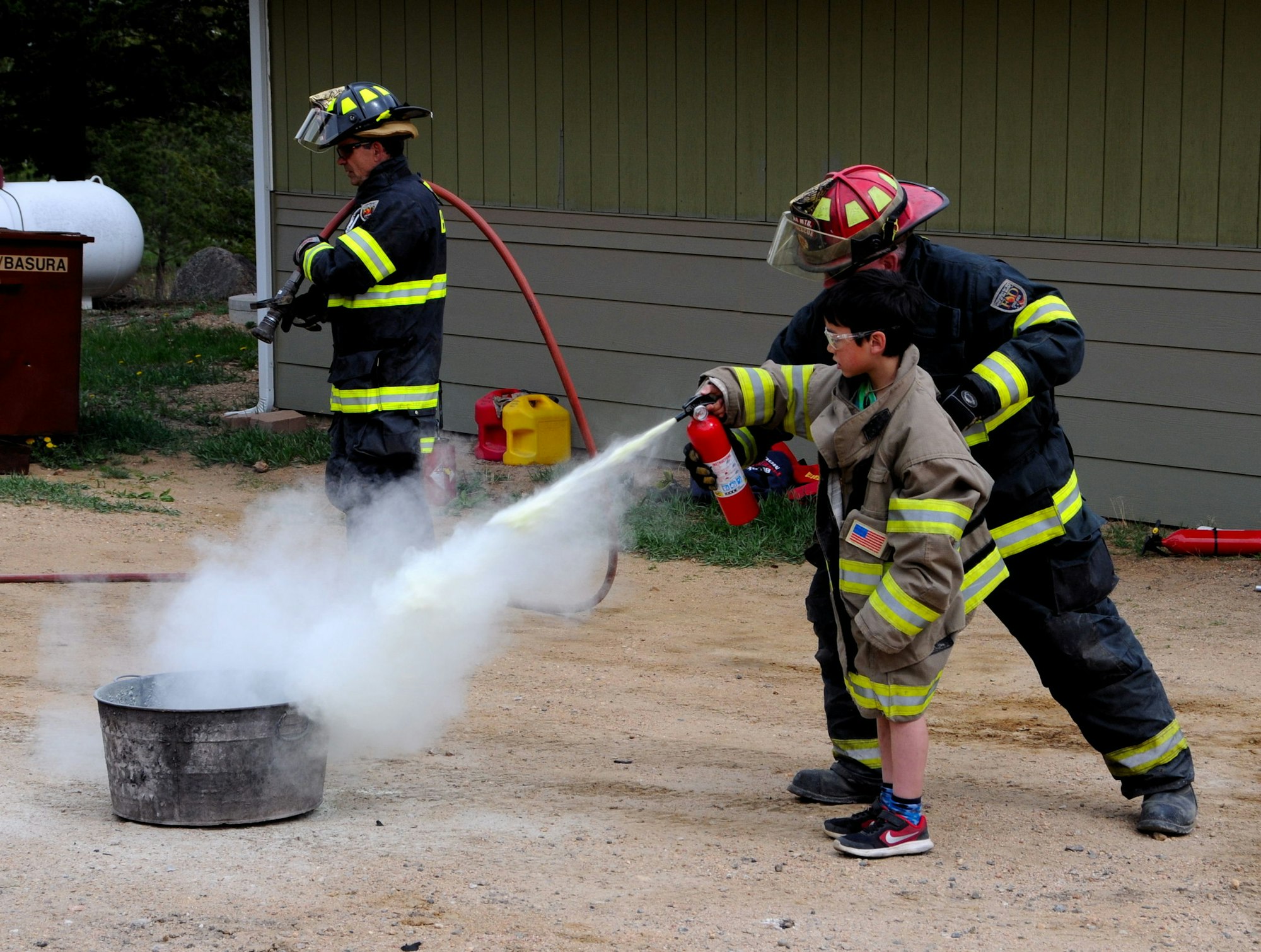 Firefighters assist a child in using a fire extinguisher on a smoky metal container.