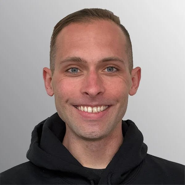 A person smiling, wearing a black hoodie against a gray background.