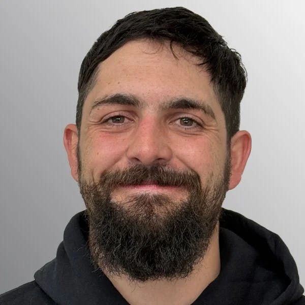 A person with short dark hair and a beard, smiling at the camera, against a gray background.
