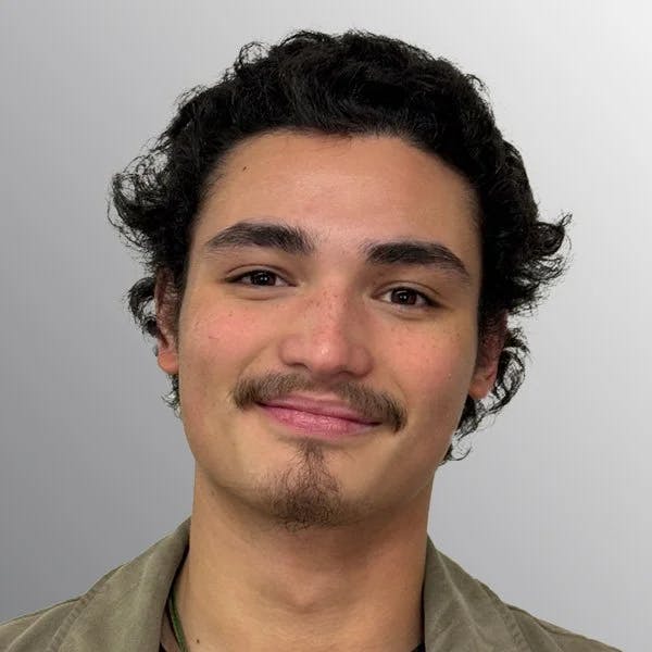 A person with curly hair and a mustache smiling against a gray background.