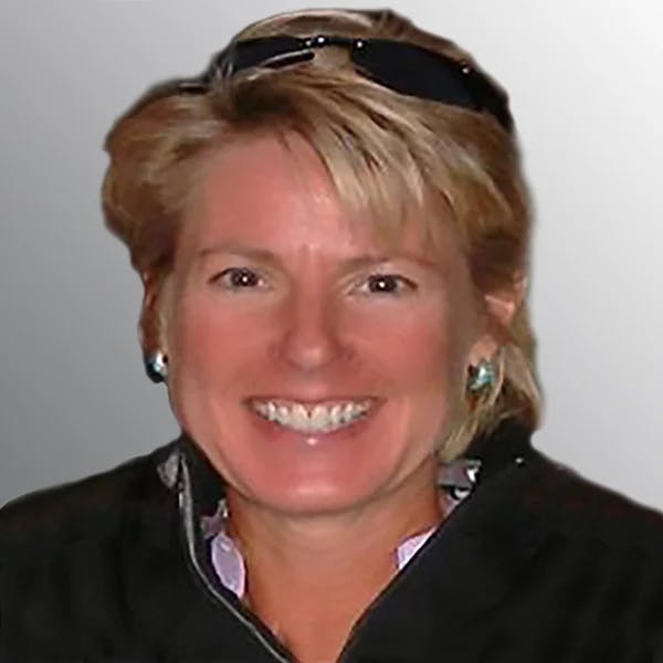 Smiling woman with short hair, wearing a black jacket, sunglasses on head, and earrings, against a gray background.