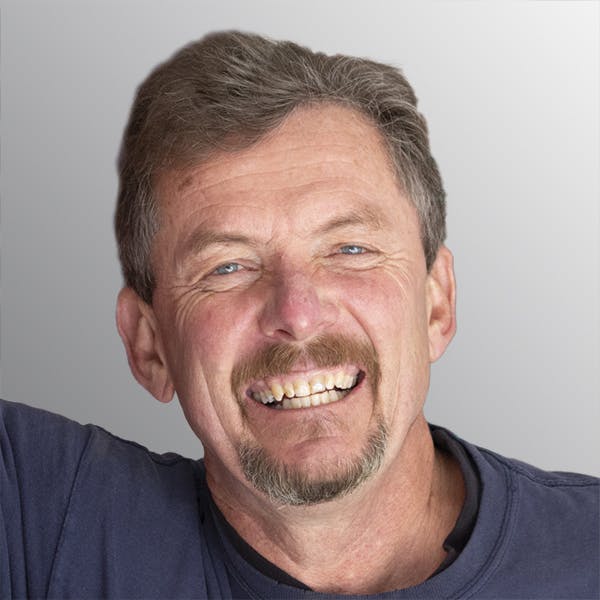 A man with a mustache and beard, smiling at the camera against a light gray background.