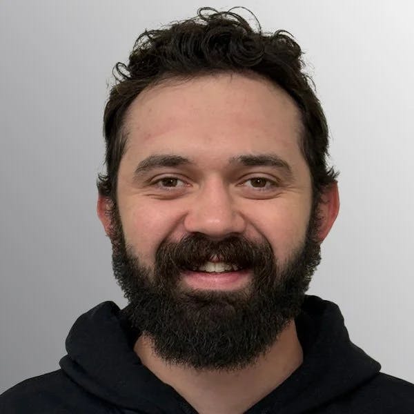 A person with dark hair and a beard, smiling, wearing a black hoodie against a neutral background.