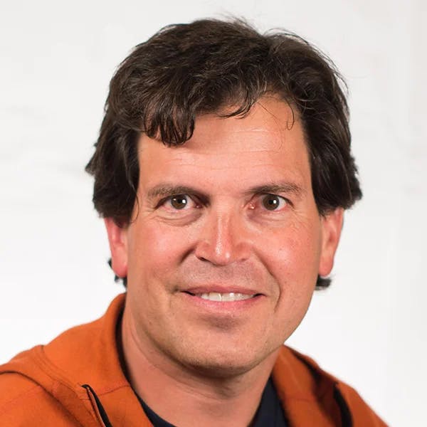 A person with dark hair, wearing an orange hoodie, smiling at the camera against a neutral background.