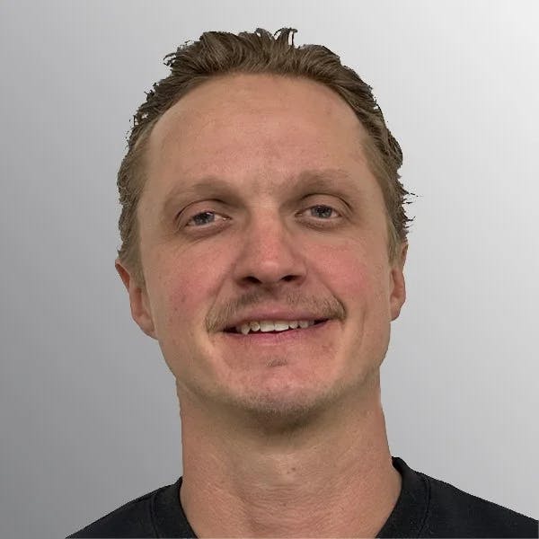 A smiling person with short hair and a light mustache against a neutral background.