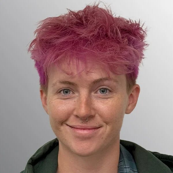 A person with short, tousled pink hair, a nose ring, and a subtle smile against a neutral background.
