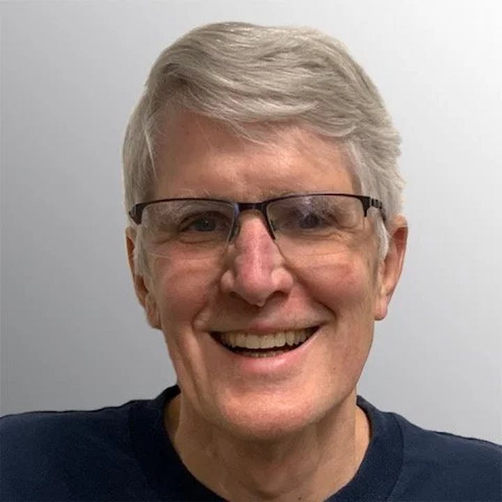 A smiling person with glasses and gray hair against a neutral background.