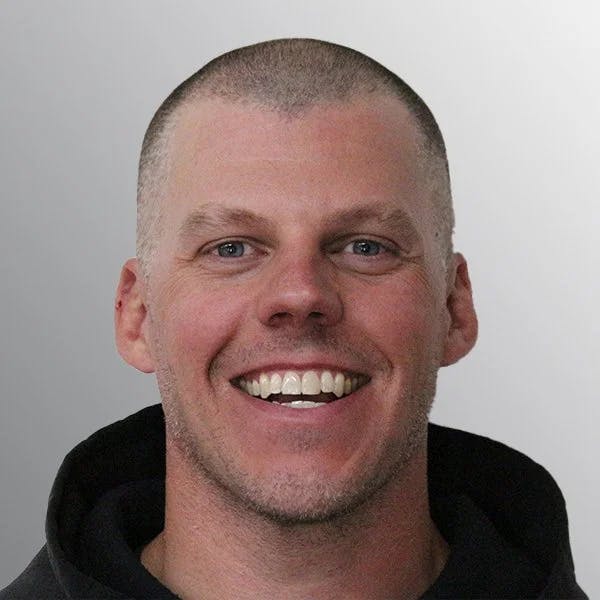 A person with a buzz cut smiling broadly against a gray background.
