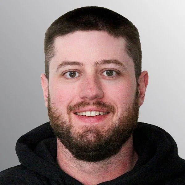 A man with short dark hair and a beard, wearing a black hoodie, smiles against a gray background.