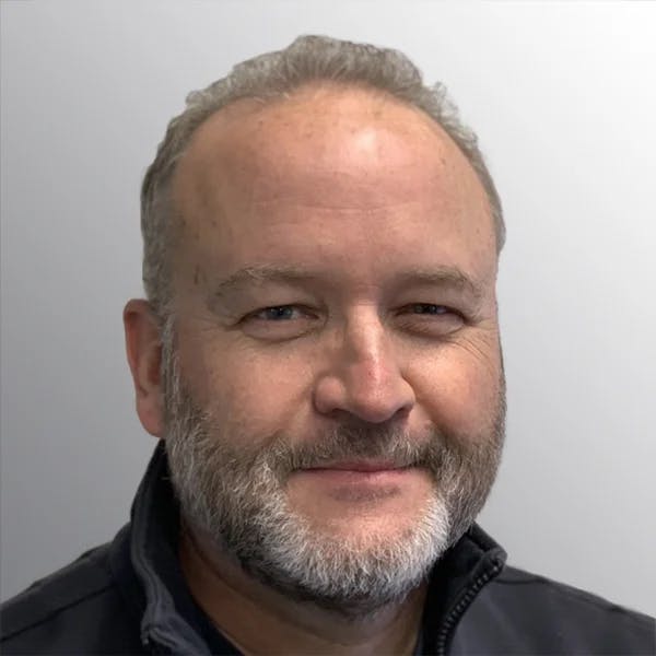 A man with short hair and a beard, wearing a dark jacket, against a neutral background.