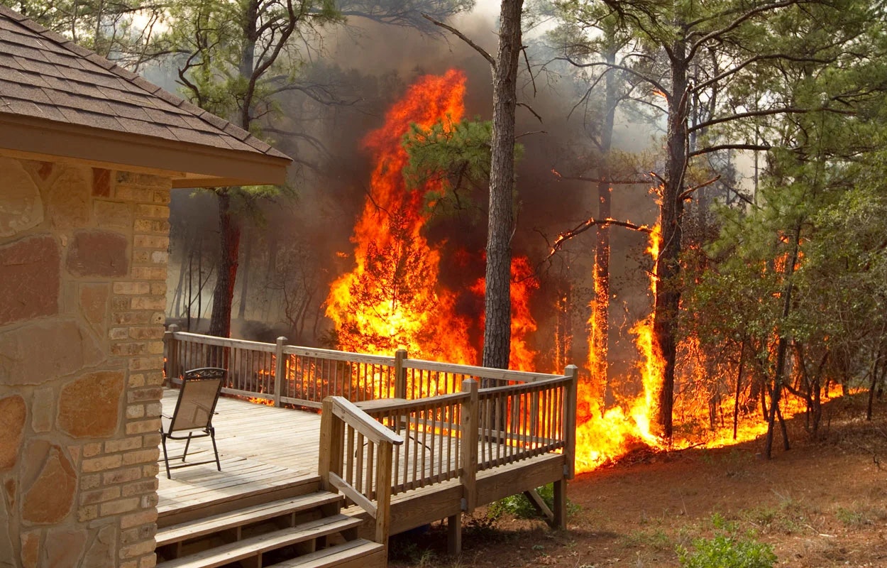 A forest fire is spreading near a wooden deck, threatening a house.