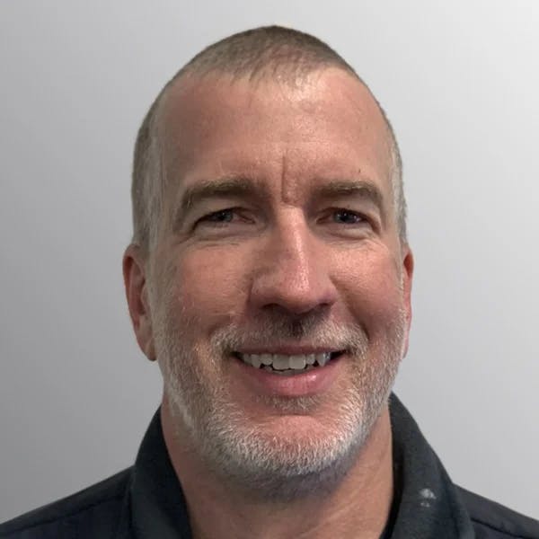 A person with short hair and a beard, smiling at the camera, against a plain background.