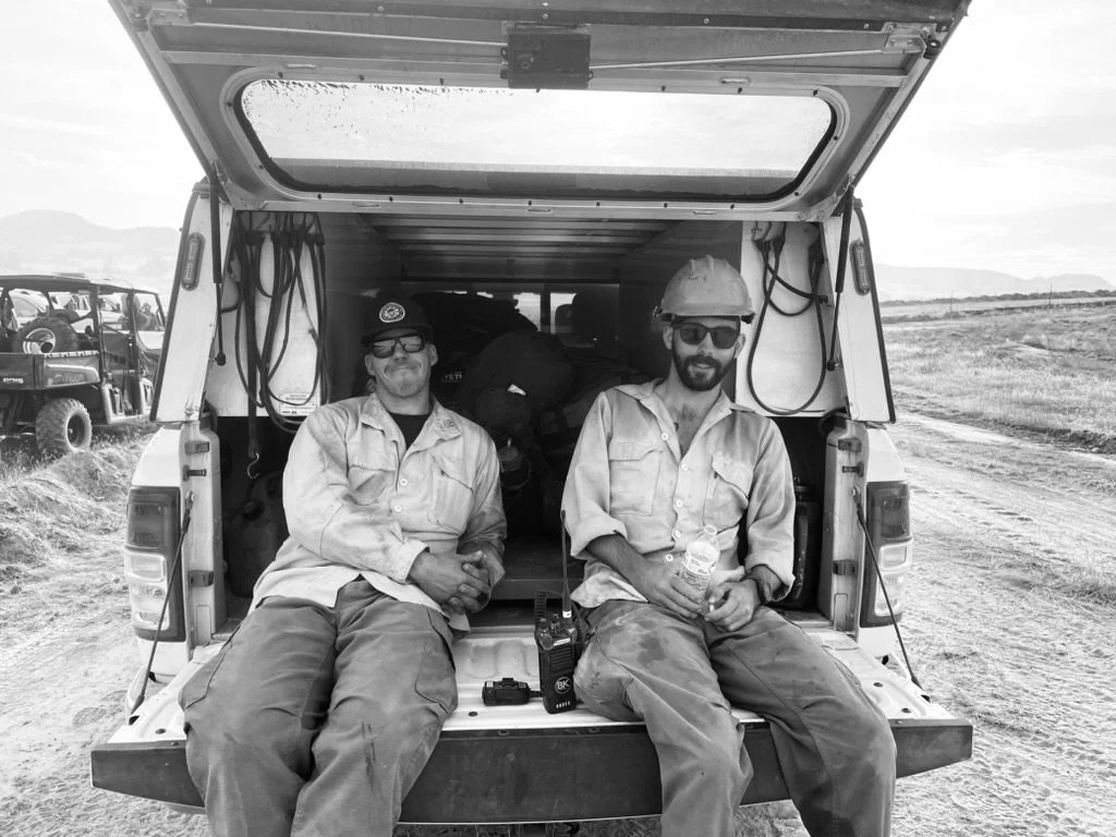 Two men in work clothes sit on the tailgate of a truck, taking a break. They’re outdoors on a dirt road with vehicles in the background.