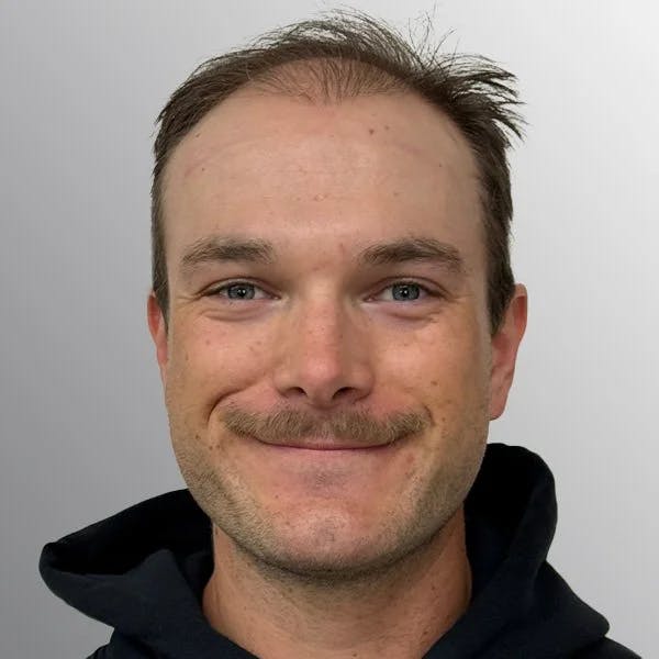 A person with a mustache smiling at the camera, wearing a dark hoodie.