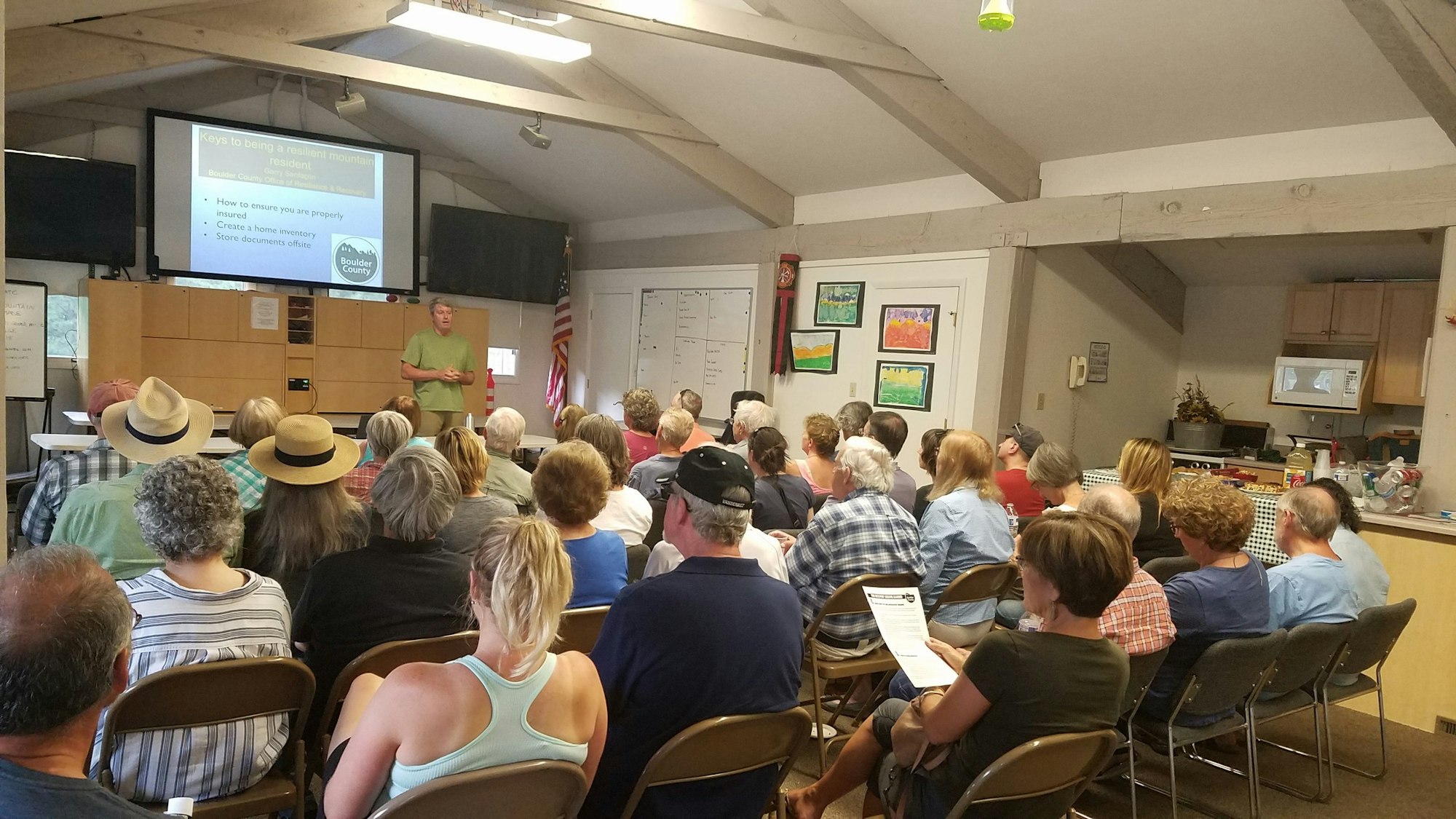 A group of people attend a presentation on being a resilient mountain resident in a room with a projection screen and U.S. flag.