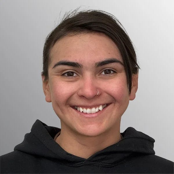 A person smiling at the camera, wearing a black hoodie and a nose ring, with short, dark hair and a light gray background.