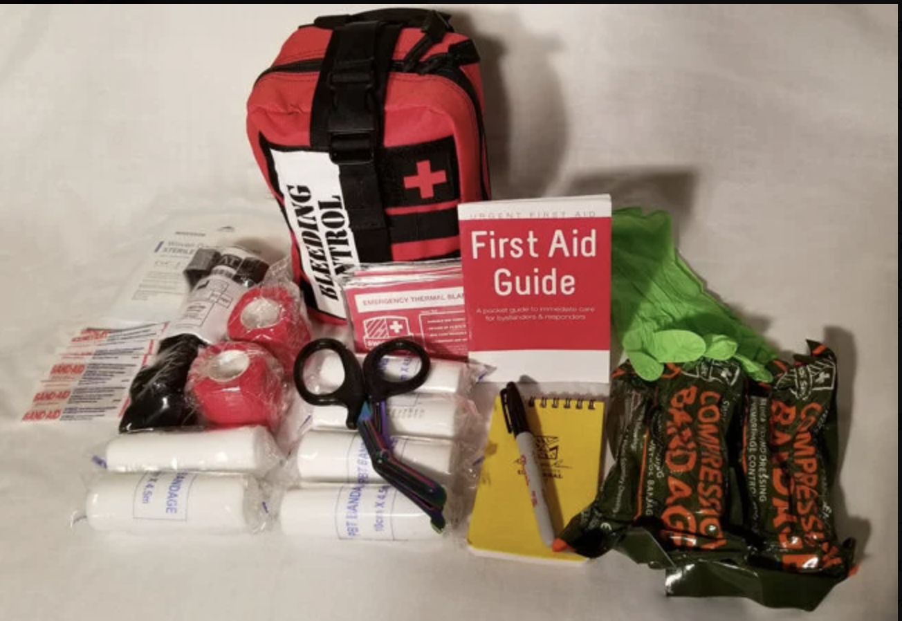 A first aid kit with a red bag, bandages, scissors, gloves, a first aid guide, notebooks, and a marker.