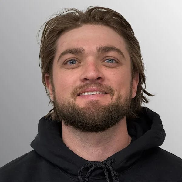 A person with a beard, wearing a black hoodie, smiling against a gray background.