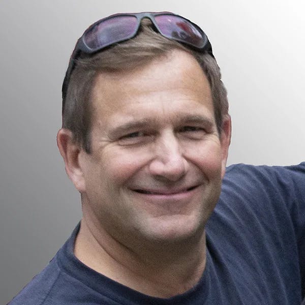 A person smiling, wearing a dark shirt and sunglasses resting on the head against a grey background.