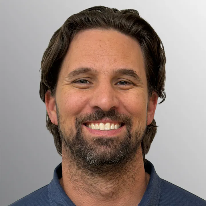 A man with brown hair and a beard, smiling, wearing a blue shirt, against a gray background.