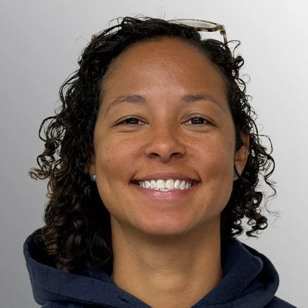 A person with curly hair, wearing glasses and a dark hoodie, smiles at the camera against a light background.