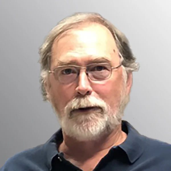 A man with glasses, light beard, and mustache, wearing a dark shirt, against a plain background.