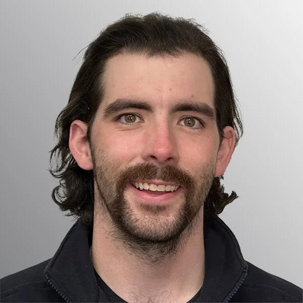 A person with dark hair, a mustache, and a goatee, wearing a dark jacket, smiles against a gray background.