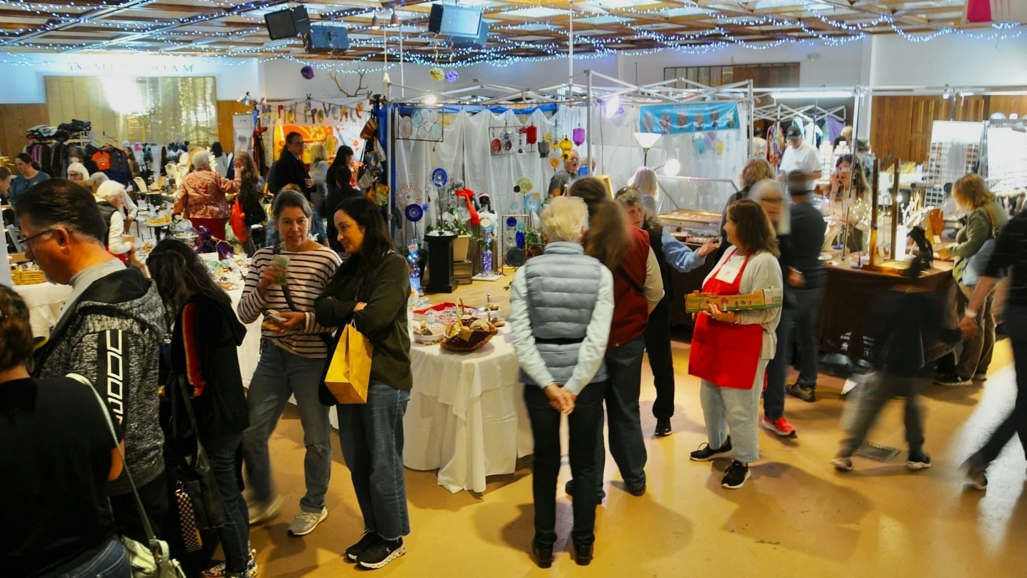 A busy indoor craft market with people browsing and interacting, stalls displaying various handmade items.