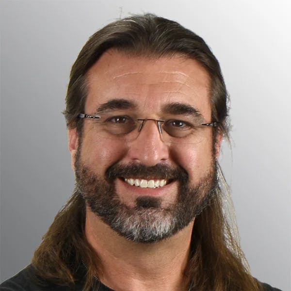 A person with long hair, a beard, and glasses, smiling against a gray background.