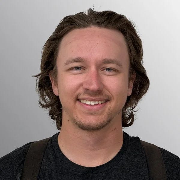 A person with long hair and a beard smiling, wearing a dark shirt and braces.