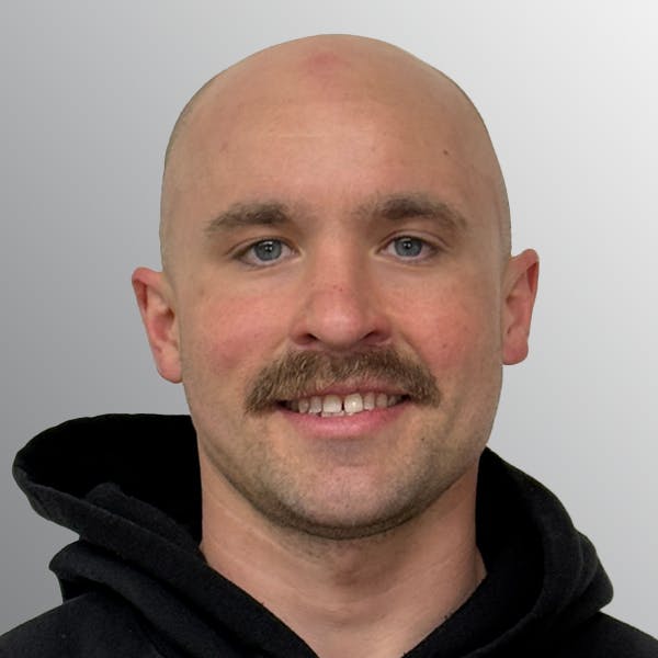 A person with a mustache and a shaved head, wearing a black hoodie, smiling at the camera against a light gray background.