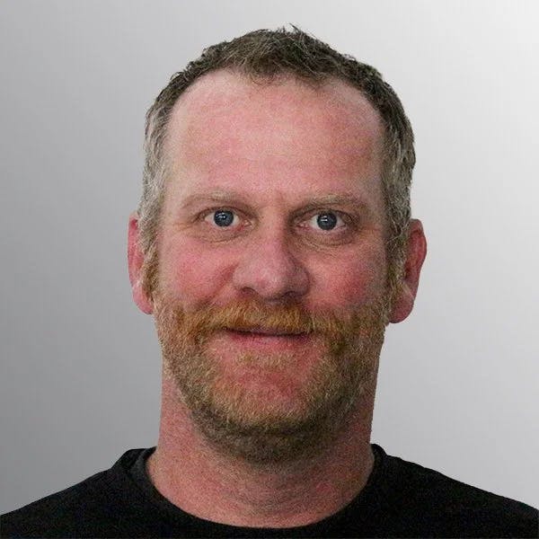 A man with short hair and a beard, wearing a black shirt, smiling at the camera against a gray background.