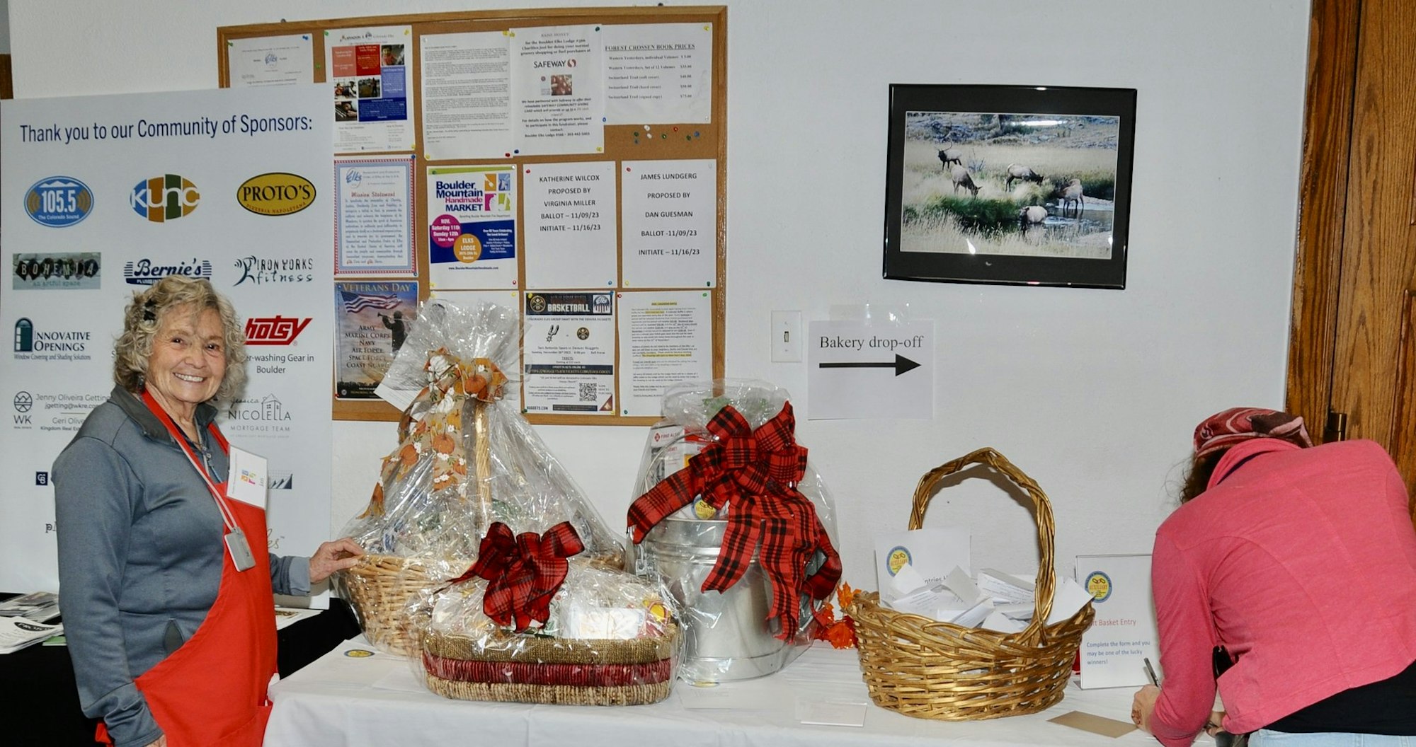 A woman in a red apron smiles next to raffle baskets. Another person is writing near a "Bakery drop-off" sign. A sponsor board is visible.