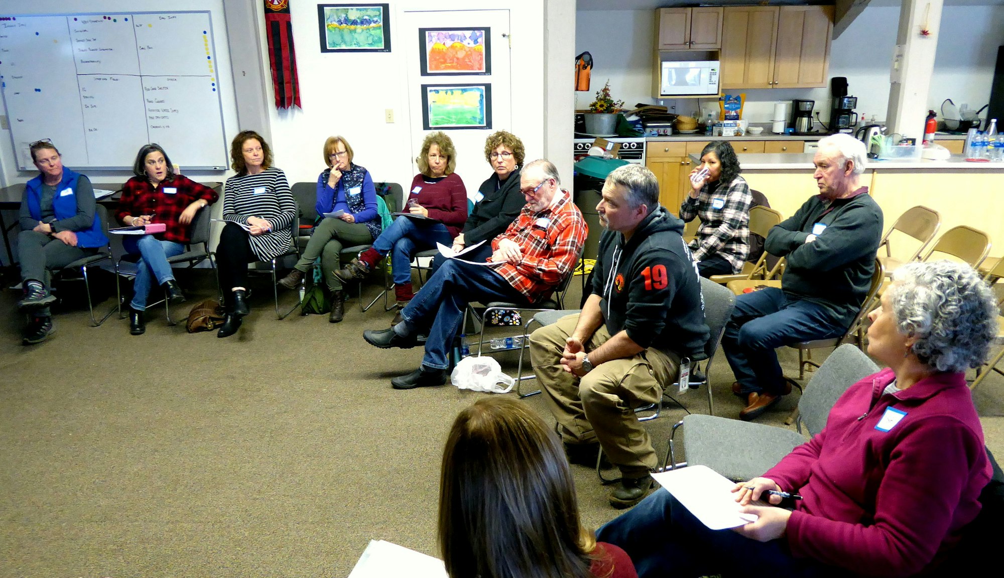 A group of people sitting in a circle having a discussion in a room with a whiteboard and kitchen in the background.