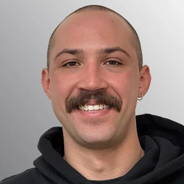 A person with a mustache, earring, and a shaved head wearing a black hoodie, smiling at the camera against a gray background.