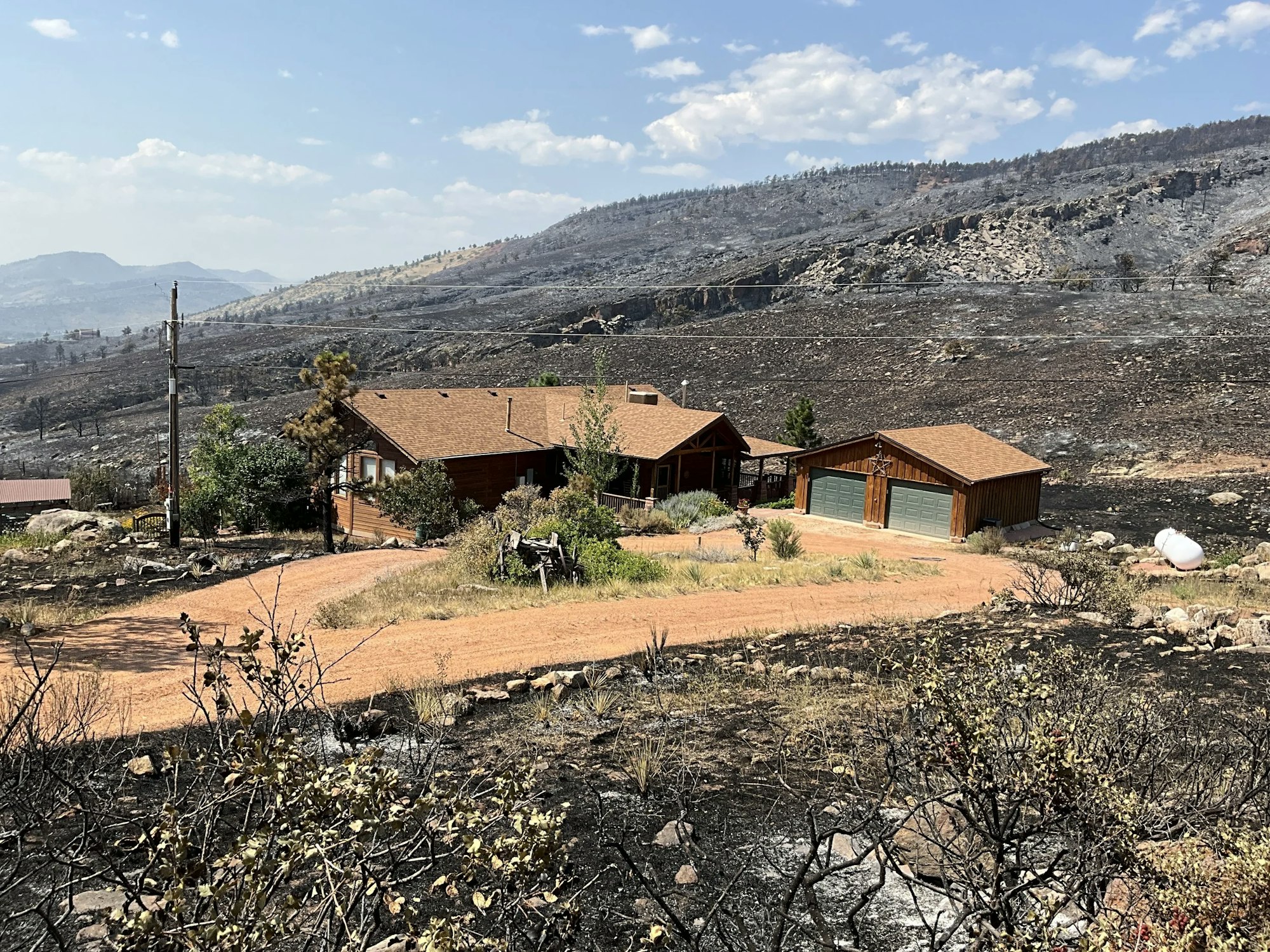 A house with a garage stands amidst a landscape of burnt hills and vegetation, indicating recent fire damage.