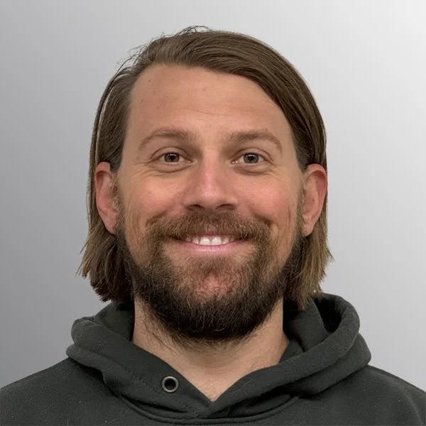 A person with a beard and long hair, wearing a black hoodie, smiling against a gray background.