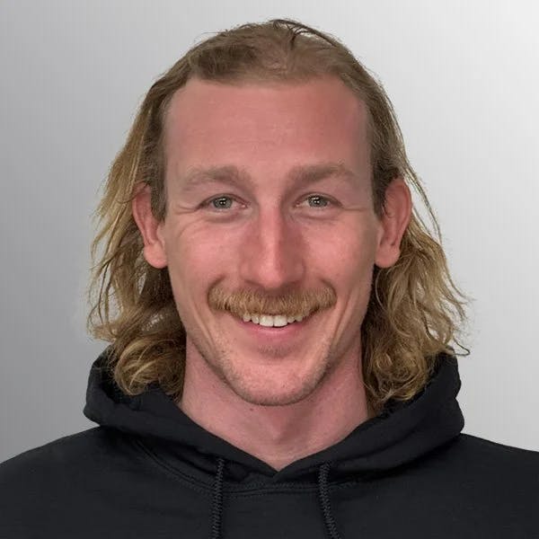 A person with long hair and a mustache smiling, wearing a black hoodie against a gray background.