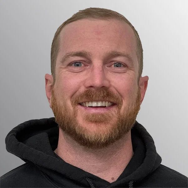 A person with a beard, smiling, wearing a black hoodie against a gray background.