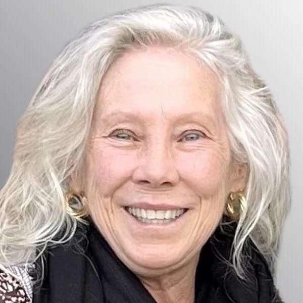 An older adult with white hair, smiling, wearing earrings and a dark scarf or sweater.