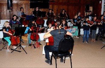 May contain: indoors, person, adult, male, man, chair, furniture, clothing, footwear, shoe, female, woman, musical instrument, leisure activities, music, musician, performer, electronics, speaker, cello, orchestra, room, accessories, jewelry, and necklace