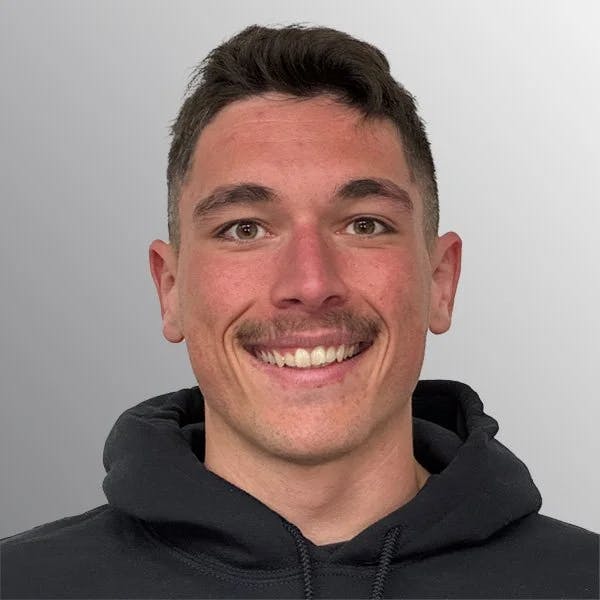 A person with short hair and a mustache, smiling, wearing a black hoodie against a gray background.