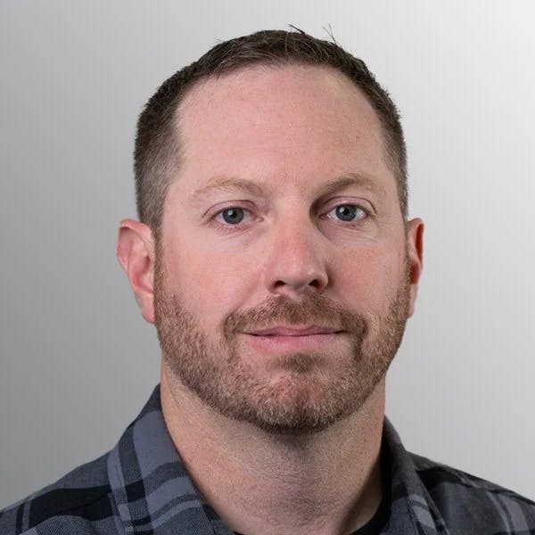 A person with short hair and a beard, wearing a checkered shirt, against a gray background.